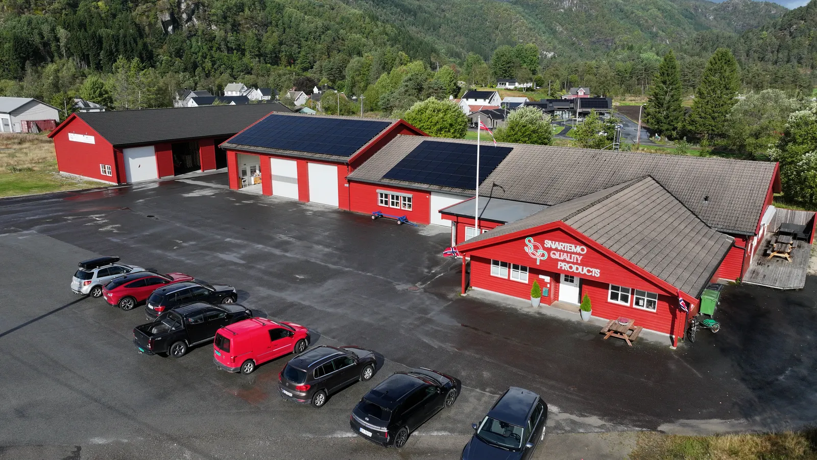 Dronebilde av SQPs røde bygninger på Snartemo med solcellepaneler på taket, parkerte biler og grønne åser i bakgrunnen