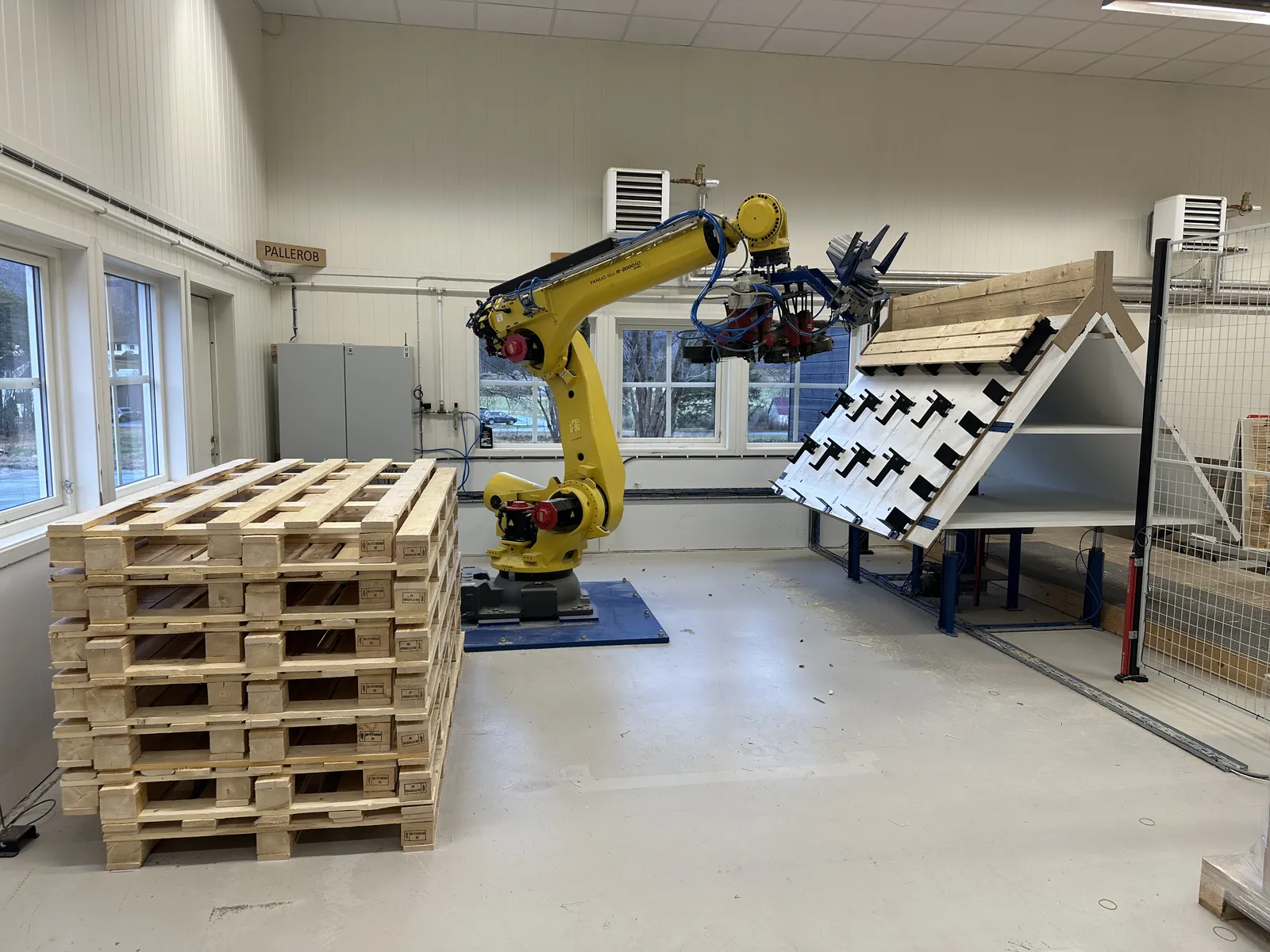 Gul industrirobot som setter sammen paller i SQPs produksjonshall, med en stabel ferdige trepaller i forgrunnen